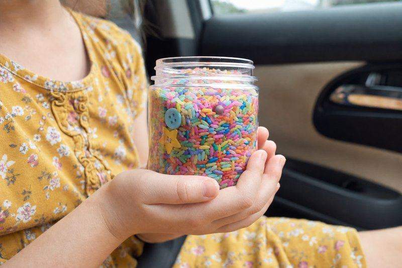 A clear jar filled with colorful rice and small hidden trinkets, held in a child's hands in a car — AI-generated image