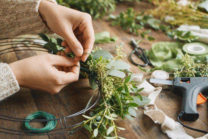 Hands using floral wire to attach greenery sprigs onto a wire wreath frame at a craft table — AI-generated image