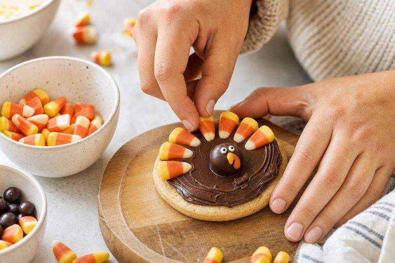 Hands pressing candy corn pieces into chocolate icing on a round cookie to form turkey feathers — AI-generated image