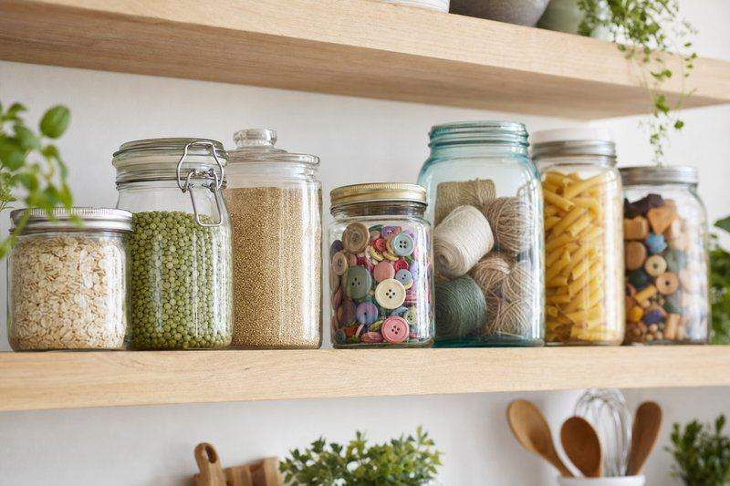 A row of mismatched glass jars on a kitchen shelf, reused to hold pantry goods and craft supplies — AI-generated image