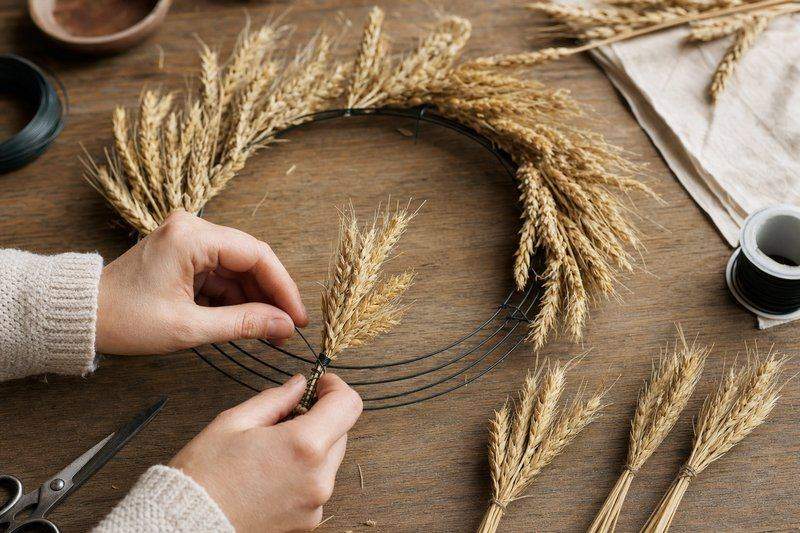 Hands wiring small bundles of dried wheat stalks onto a wire wreath form, mid-construction — AI-generated image
