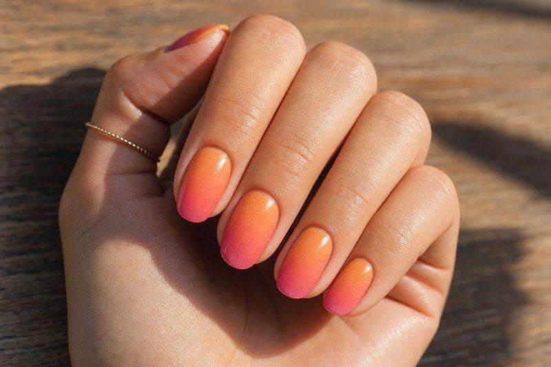 A hand with a sunset-style ombre manicure blending orange into pink across the nails — AI-generated image