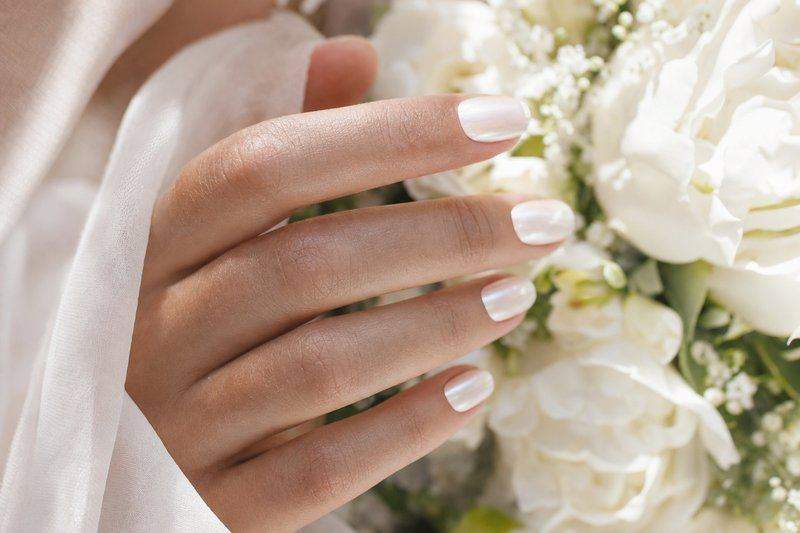 A hand with a soft milky-white pearlescent nail polish finish, resting near white wedding flowers — AI-generated image